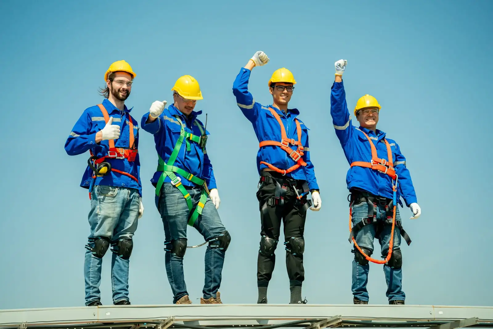 Grup de lucrători cu căști galbene și hamuri, pe un acoperiș, sub cer senin